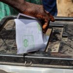Mão deposita uma cédula de papel em uma urna eleitoral transparente ao ar livre.