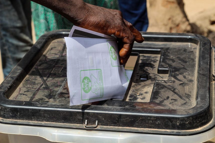 Mão deposita uma cédula de papel em uma urna eleitoral transparente ao ar livre.