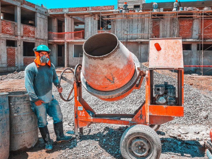 Trabalhador da construção civil opera uma betoneira num estaleiro de obras, usando equipamento de proteção.