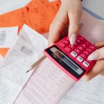 Mãos usando uma calculadora rosa entre recibos e documentos financeiros sobre uma mesa.