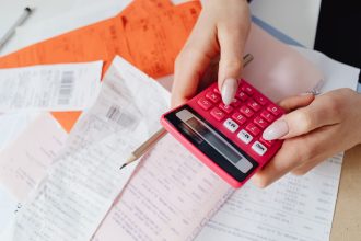 Mãos usando uma calculadora rosa entre recibos e documentos financeiros sobre uma mesa.