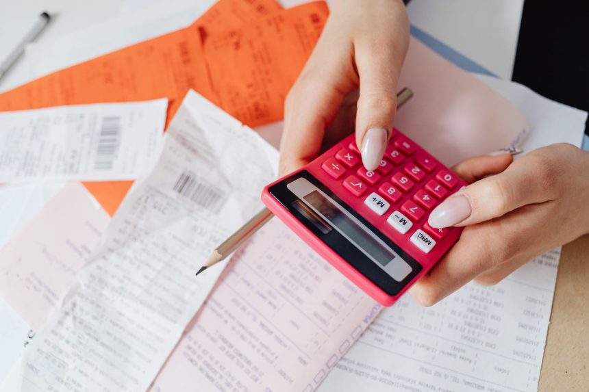 Mãos usando uma calculadora rosa entre recibos e documentos financeiros sobre uma mesa.