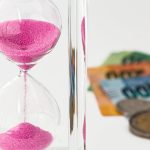 Ampulheta sobre moedas e cédulas, representando espera e questões financeiras.